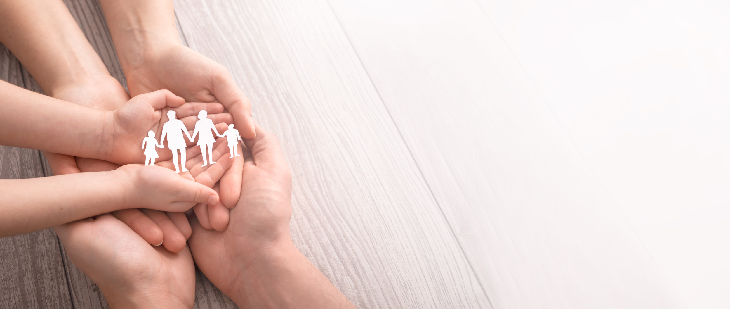 Hände von Kinder und Eltern die offen übereinander auf den Tisch liegen und eine Familie aus Papier festhalten.